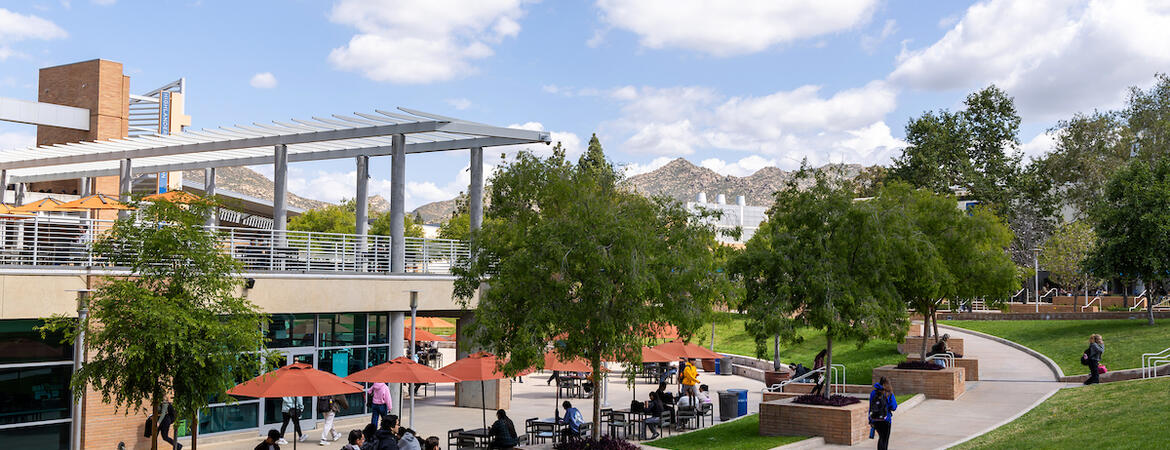 UCR campus: modern building, outdoor seating, green lawn, students, distant hills.