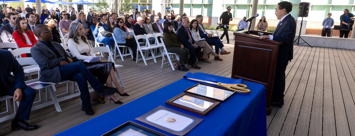 Man speaks at podium to outdoor audience; framed documents on blue table.