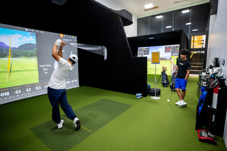 Golfer swings club in indoor simulator, another person observes.